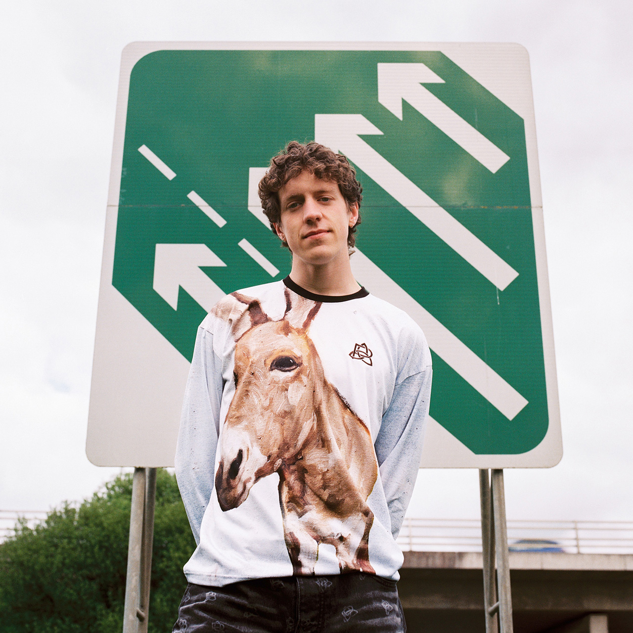 Young man wearing white graphic t-shirt with goat print standing before green exit sign in urban setting