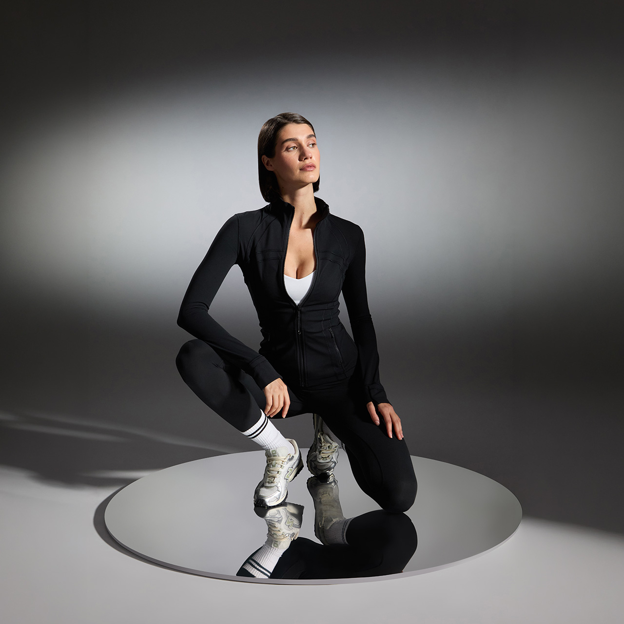 Woman in black activewear jacket and leggings wearing white trainers, crouching pose on circular platform