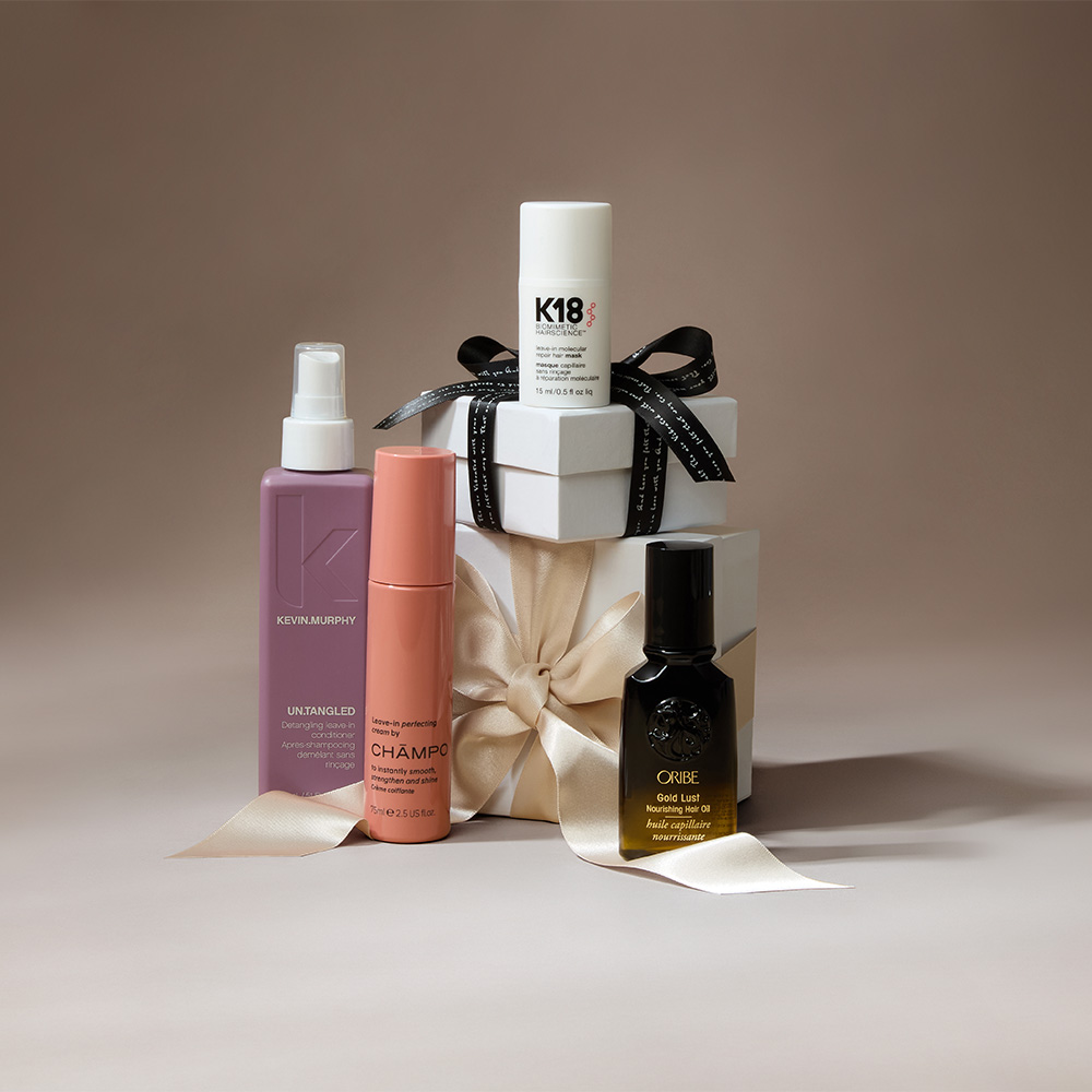 A group of hair care products arranged in front of a gift box with a ribbon on a neutral background.