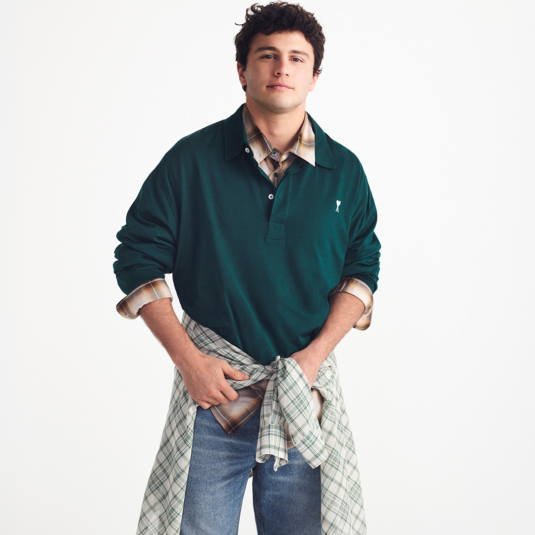Man wearing dark green polo shirt with checked shirt draped around waist and denim jeans against white background