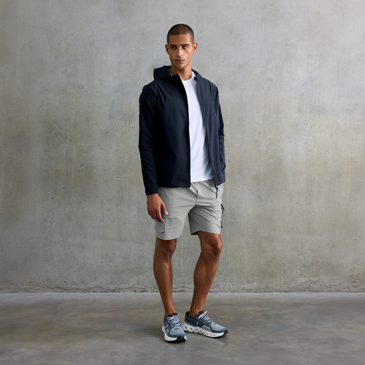 Man wearing navy zip jacket, white t-shirt and beige shorts against grey textured background