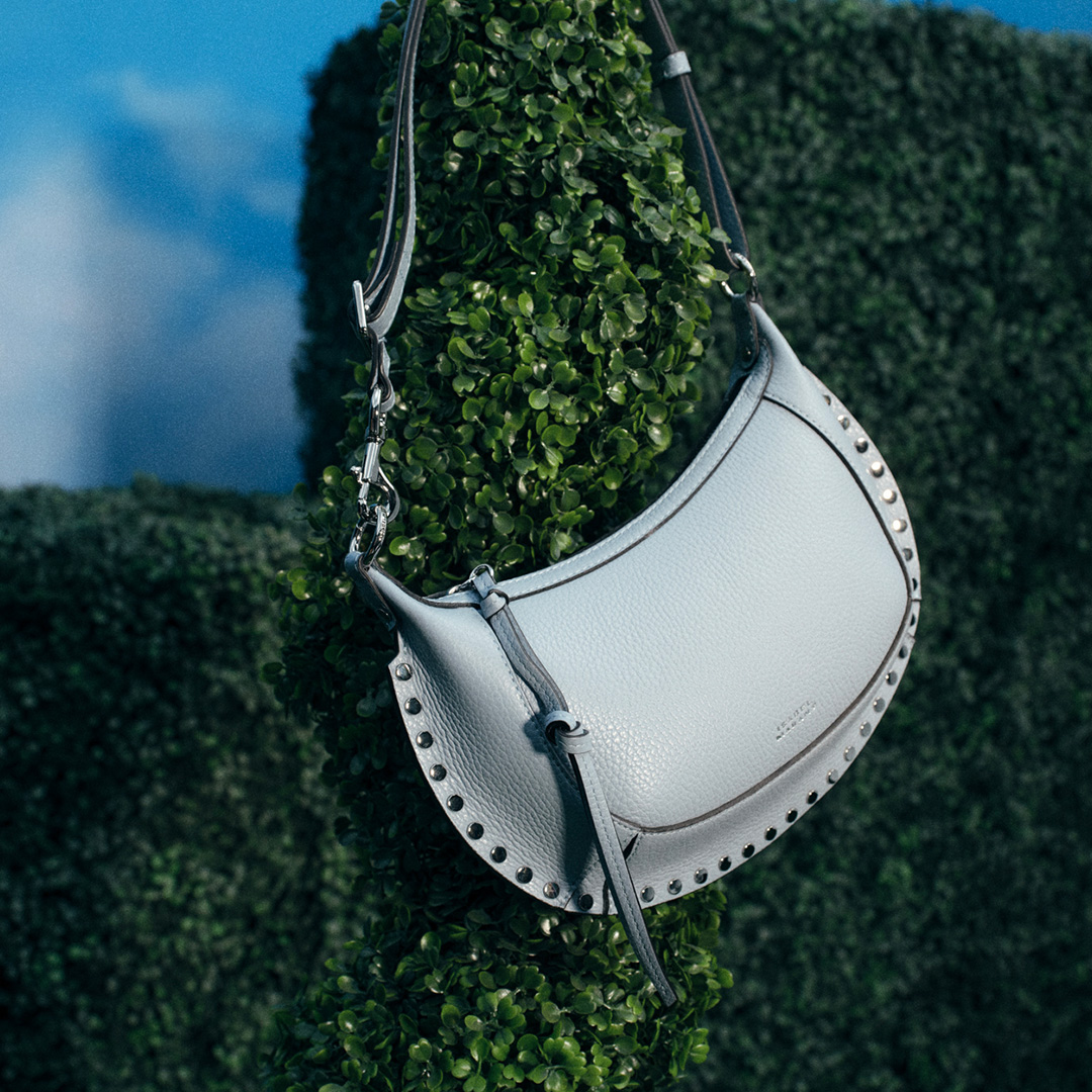 White leather shoulder bag with studded details, hanging from ivy-covered wall against blue sky