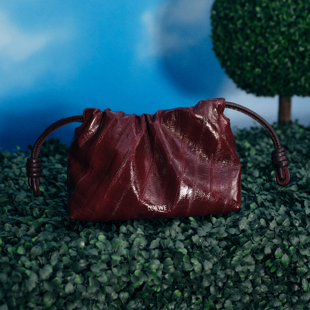 Burgundy leather crossbody bag with gathered detailing and brown handles displayed on moss ground with topiary backdrop