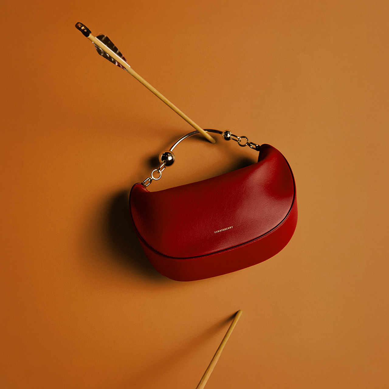 A red handbag with a metallic strap is pierced by arrows, set against an orange background.