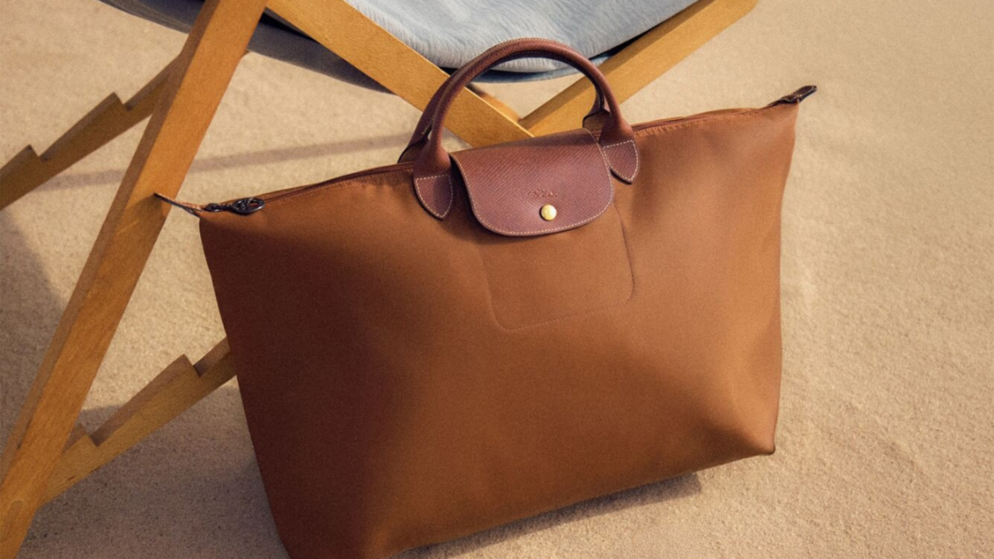 A brown leather handbag with a dark brown handle rests against a wooden chair.