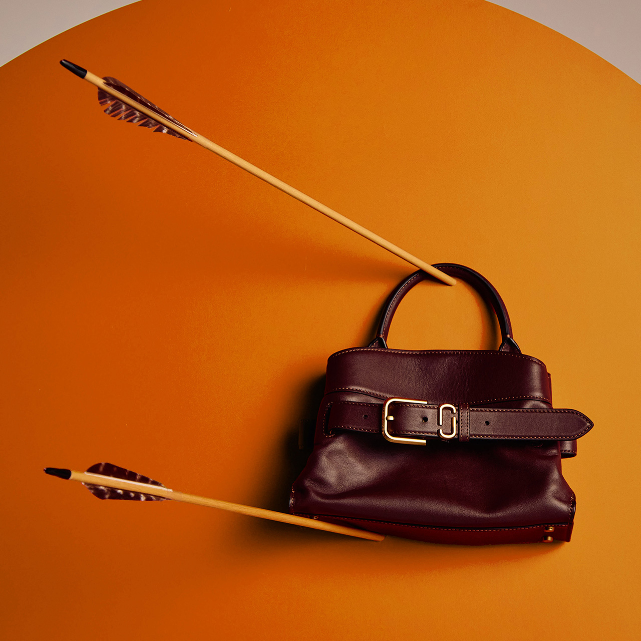 A burgundy handbag with a belt-like strap is pierced by an arrow on an orange background.