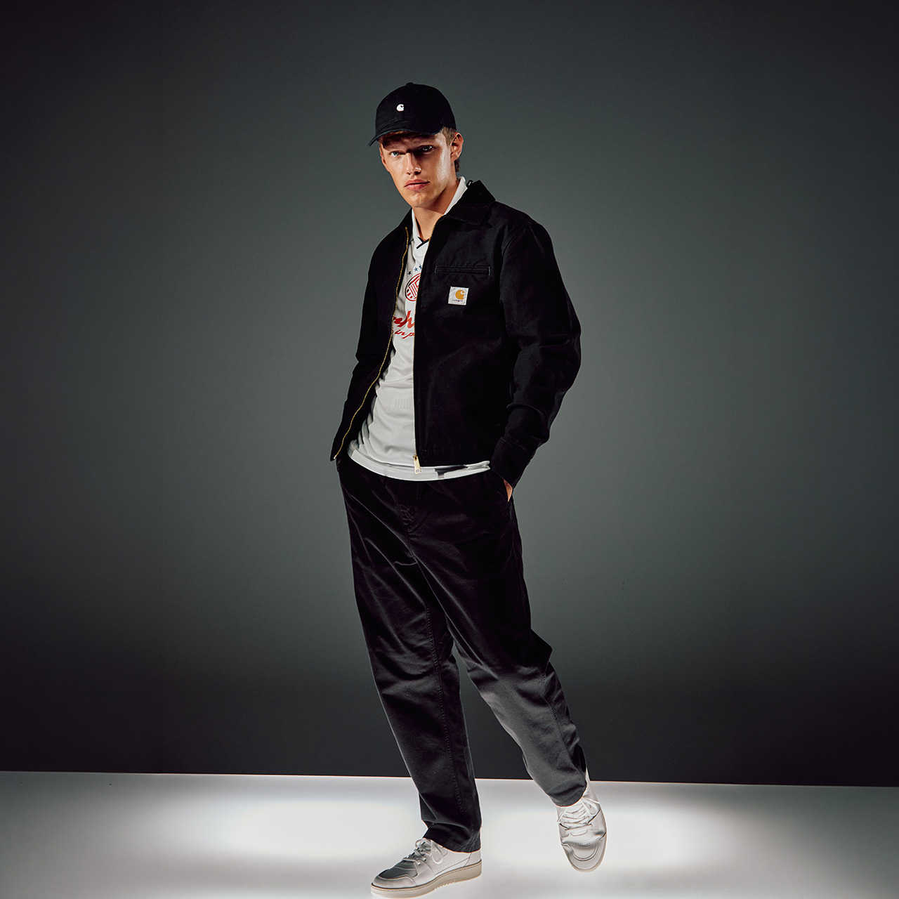 Young man in black Carhartt jacket with embroidered logo, white t-shirt, black and grey trousers, and black cap