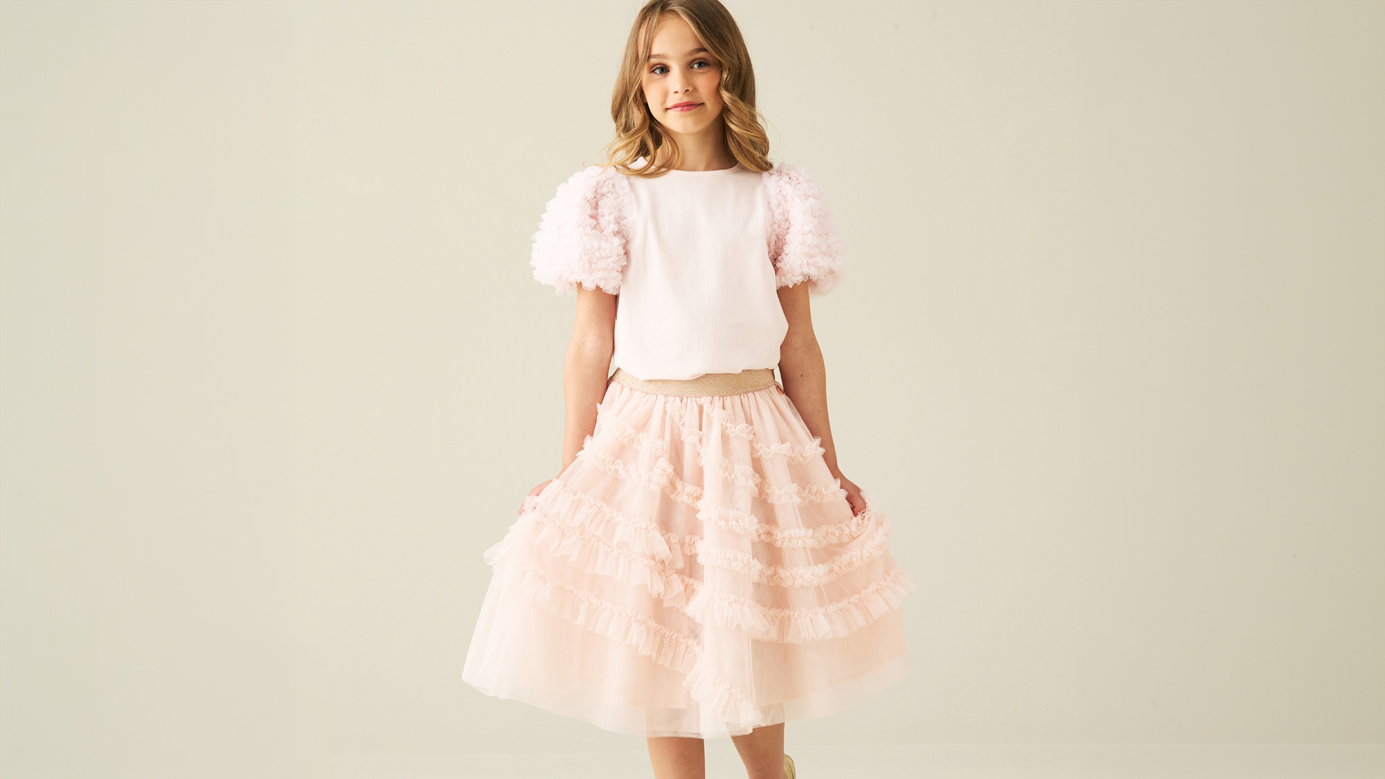 A young girl in a pink, ruffled dress and top with puffed sleeves stands against a plain background.