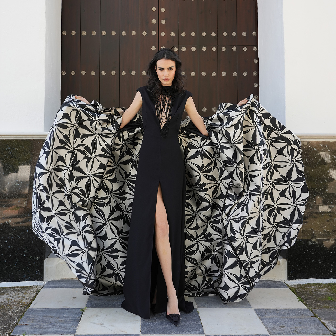 Woman in black sleeveless dress with layered gold chains standing before black and cream floral patterned fabric panels