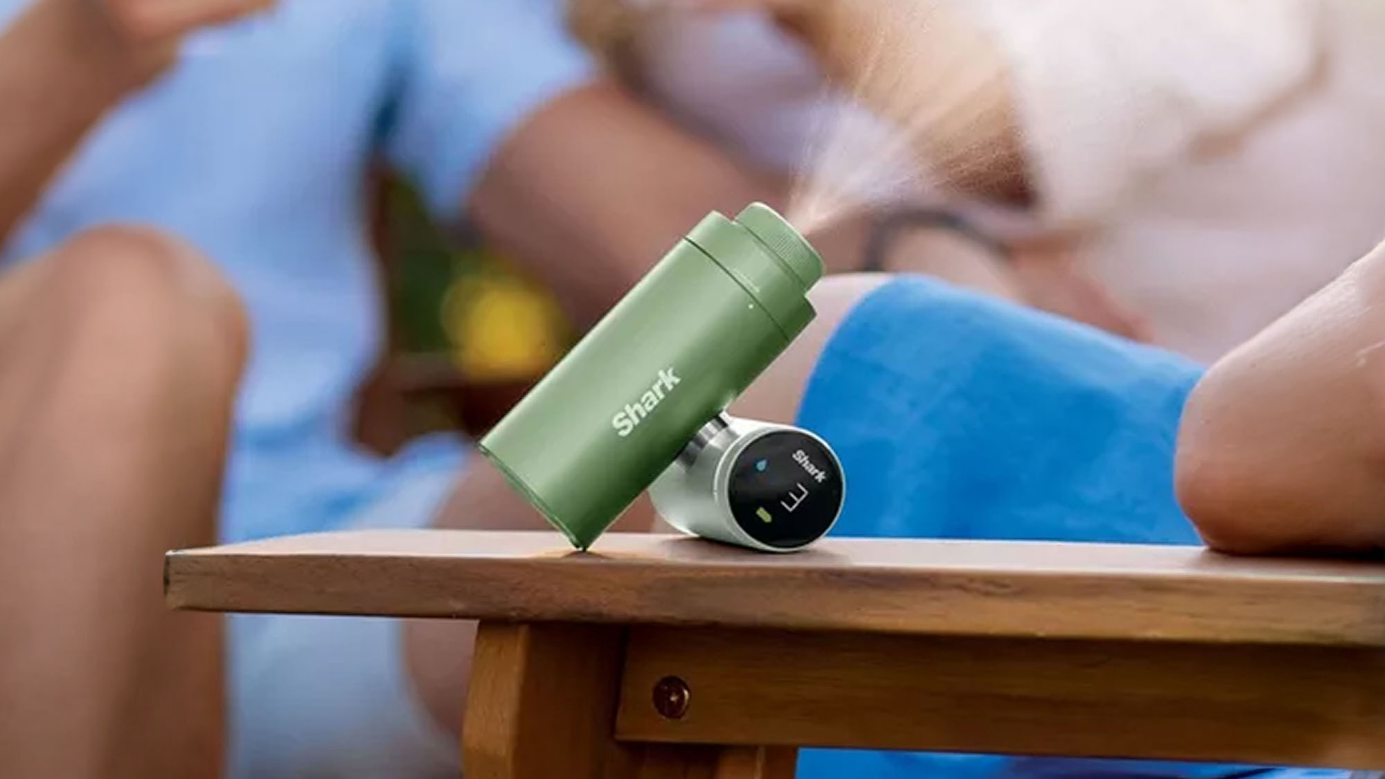 A green portable device emitting a mist is placed on a wooden outdoor table next to a person in blue attire.