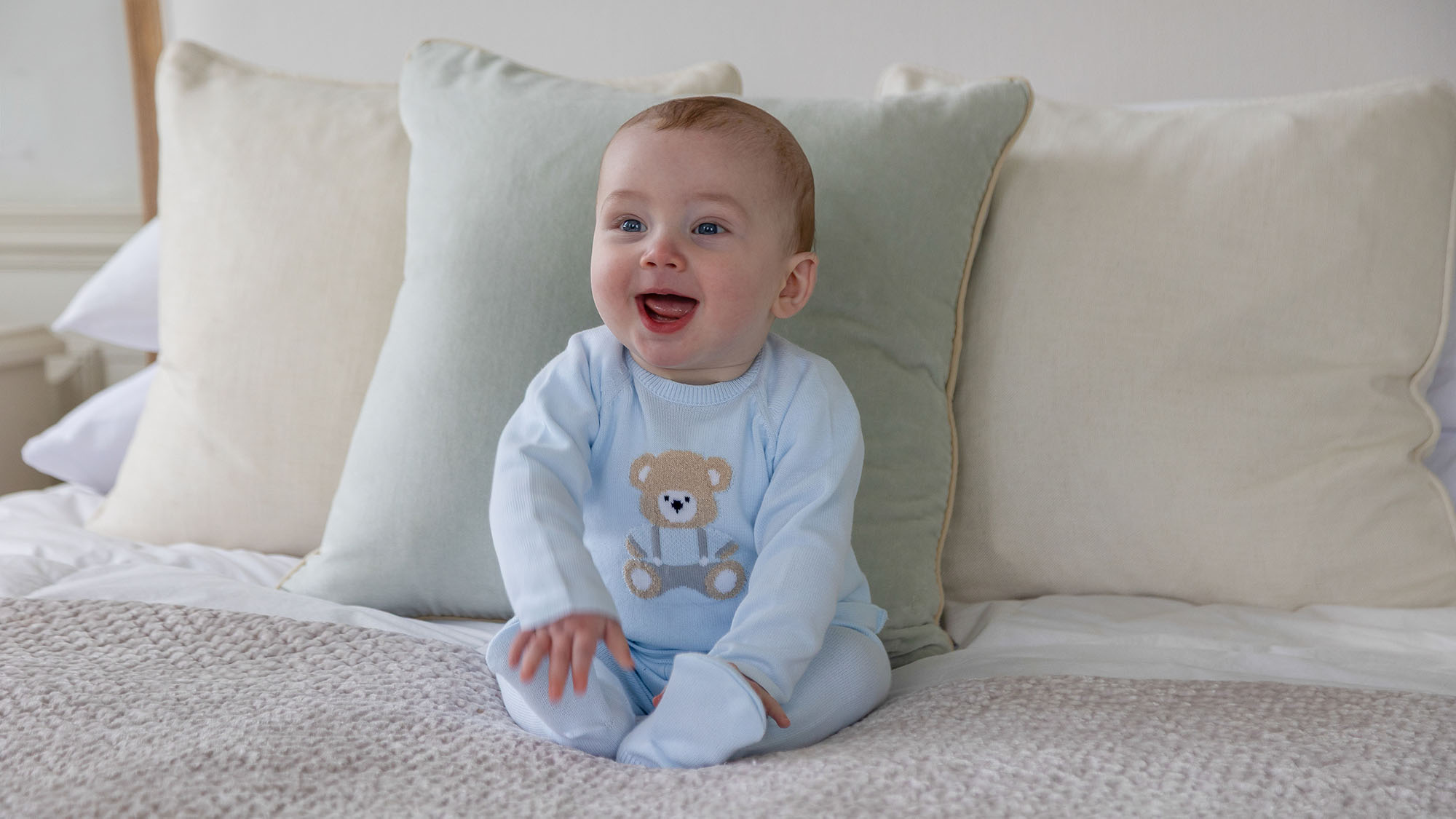 Smiling baby wearing light blue jumper with teddy bear design, sat on cream cushions
