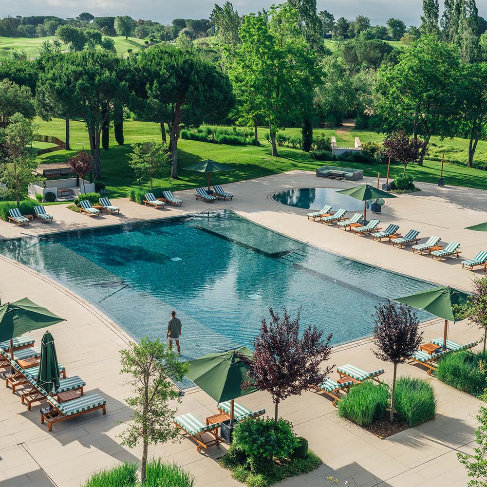 Outdoor pool area with loungers, umbrellas, and surrounding greenery on a sunny day.