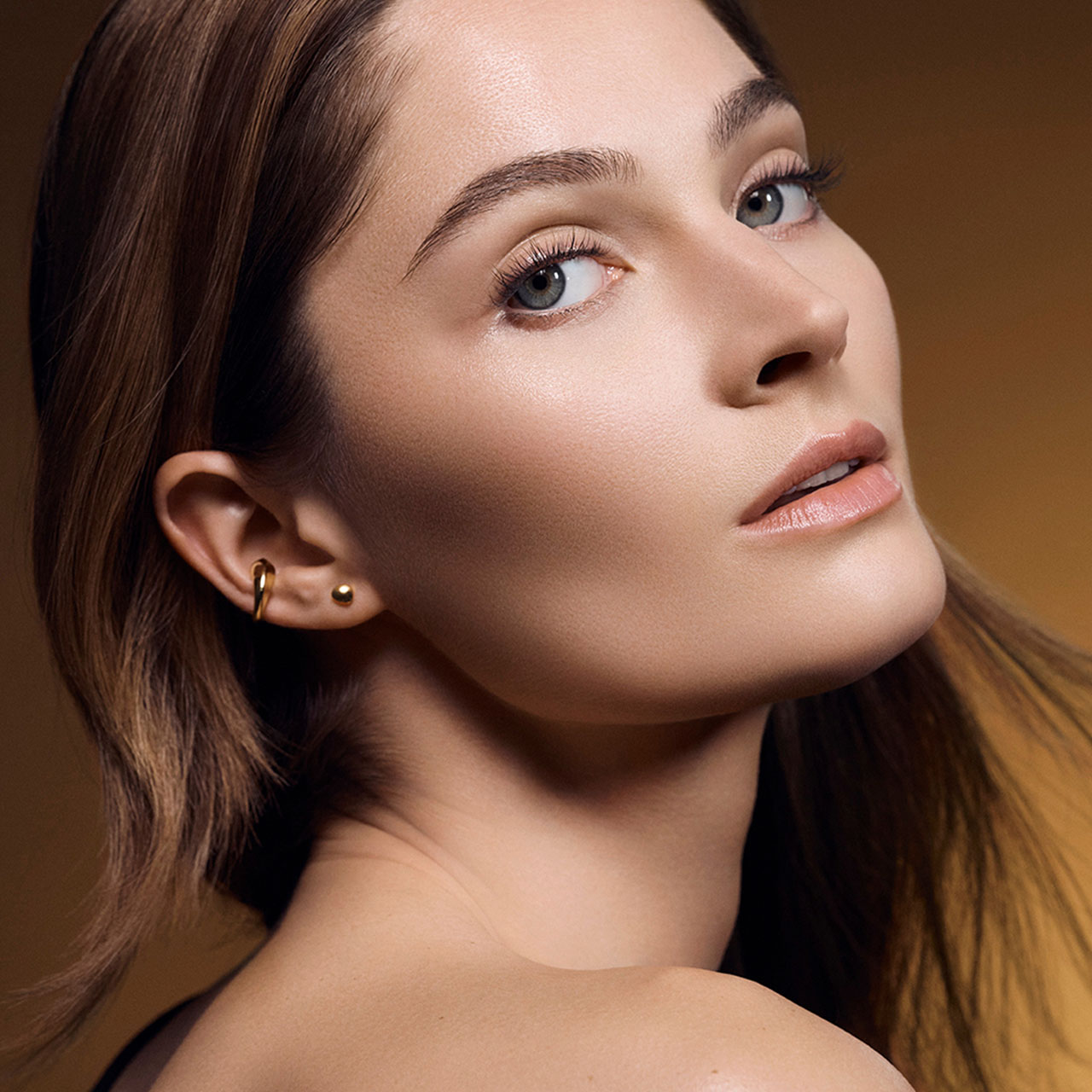 Woman with brown hair showcasing polished skin and natural makeup look against warm neutral background