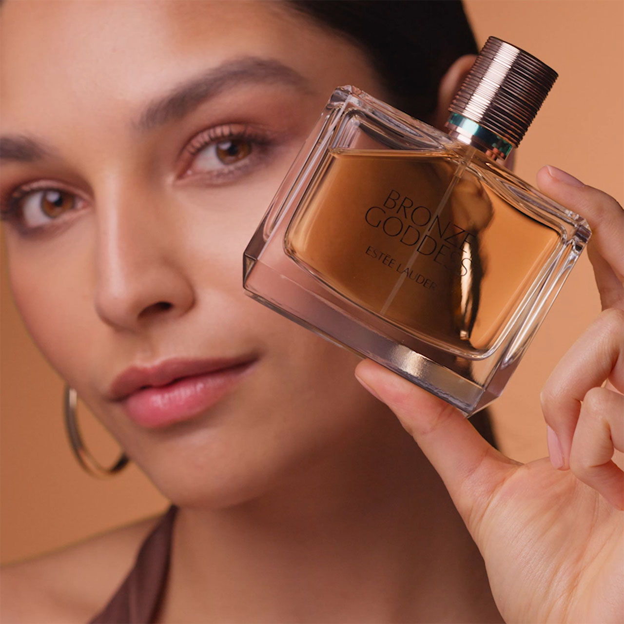 Woman holding bronze-toned fragrance bottle with metallic cap against warm peachy background