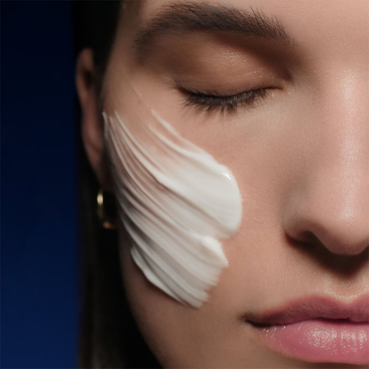 Woman with eyes closed applying white facial cream to cheek against dark blue background