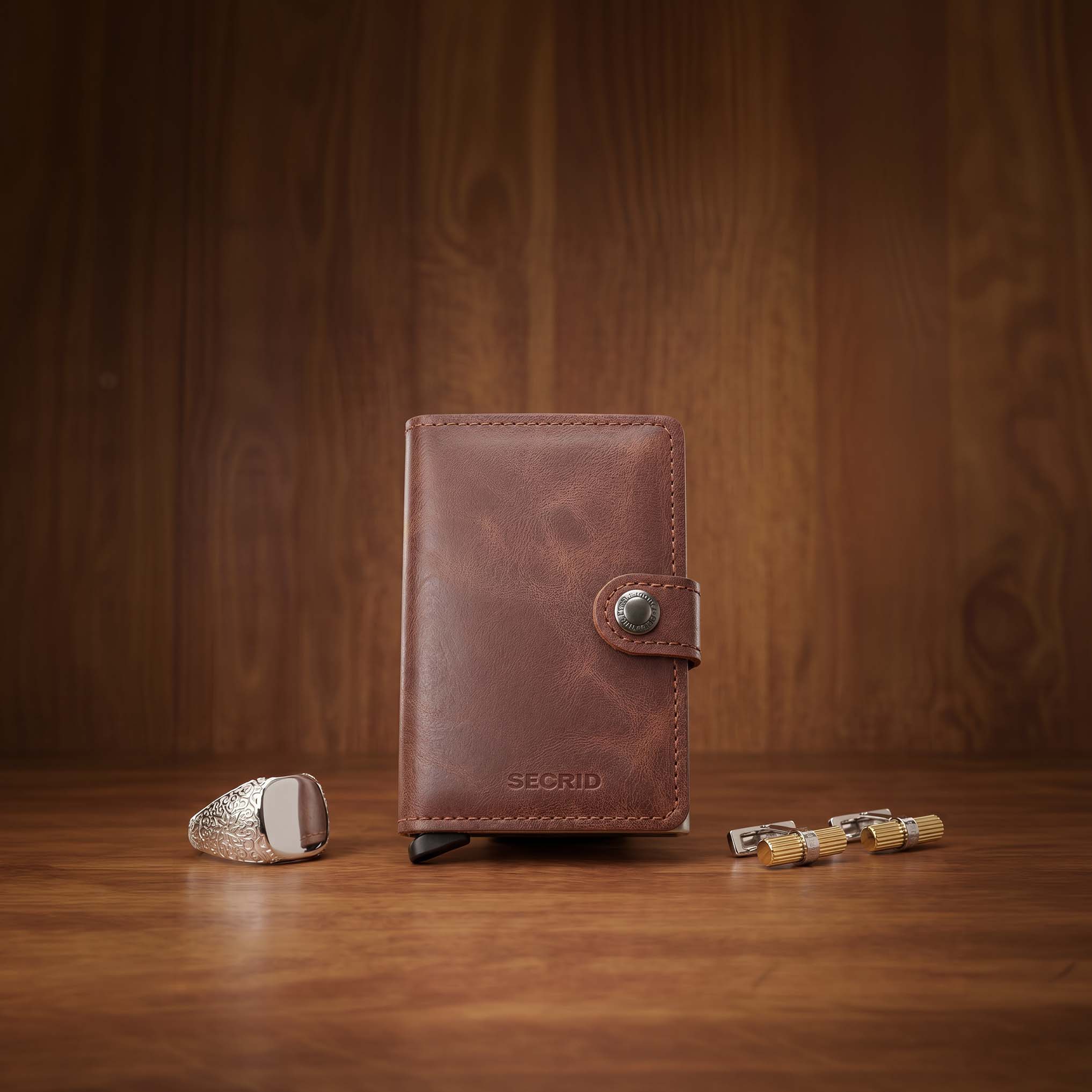 A brown leather wallet with a button clasp is placed upright on a wooden surface. Beside it, a silver ring with intricate details and a set of three small metal cylinders are arranged neatly. The background features a matching wooden pattern.