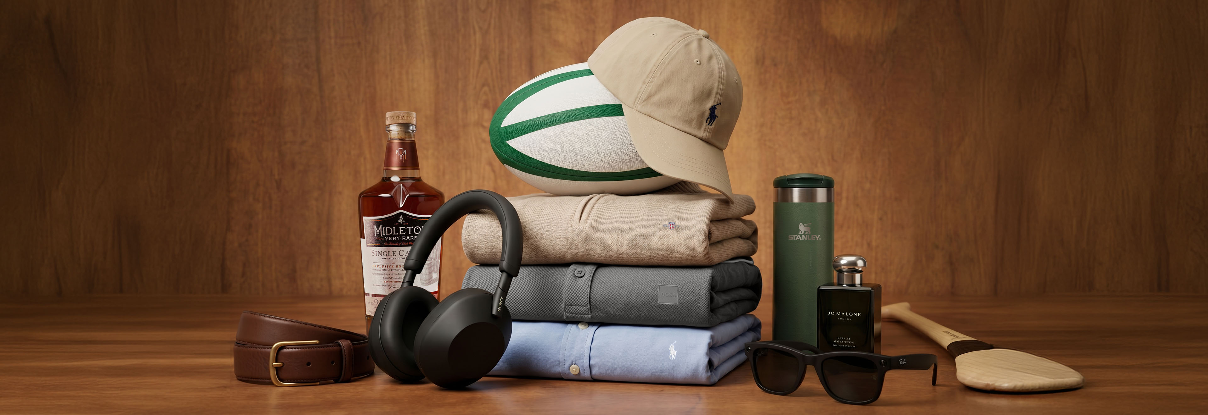 A collection of items on a wooden surface, including a beige cap, an Irish Rugby Football Union ball, a bottle of Midleton whiskey, a brown leather belt, headphones, folded shirts in neutral tones, a green water bottle, sunglasses, and men's cologne.