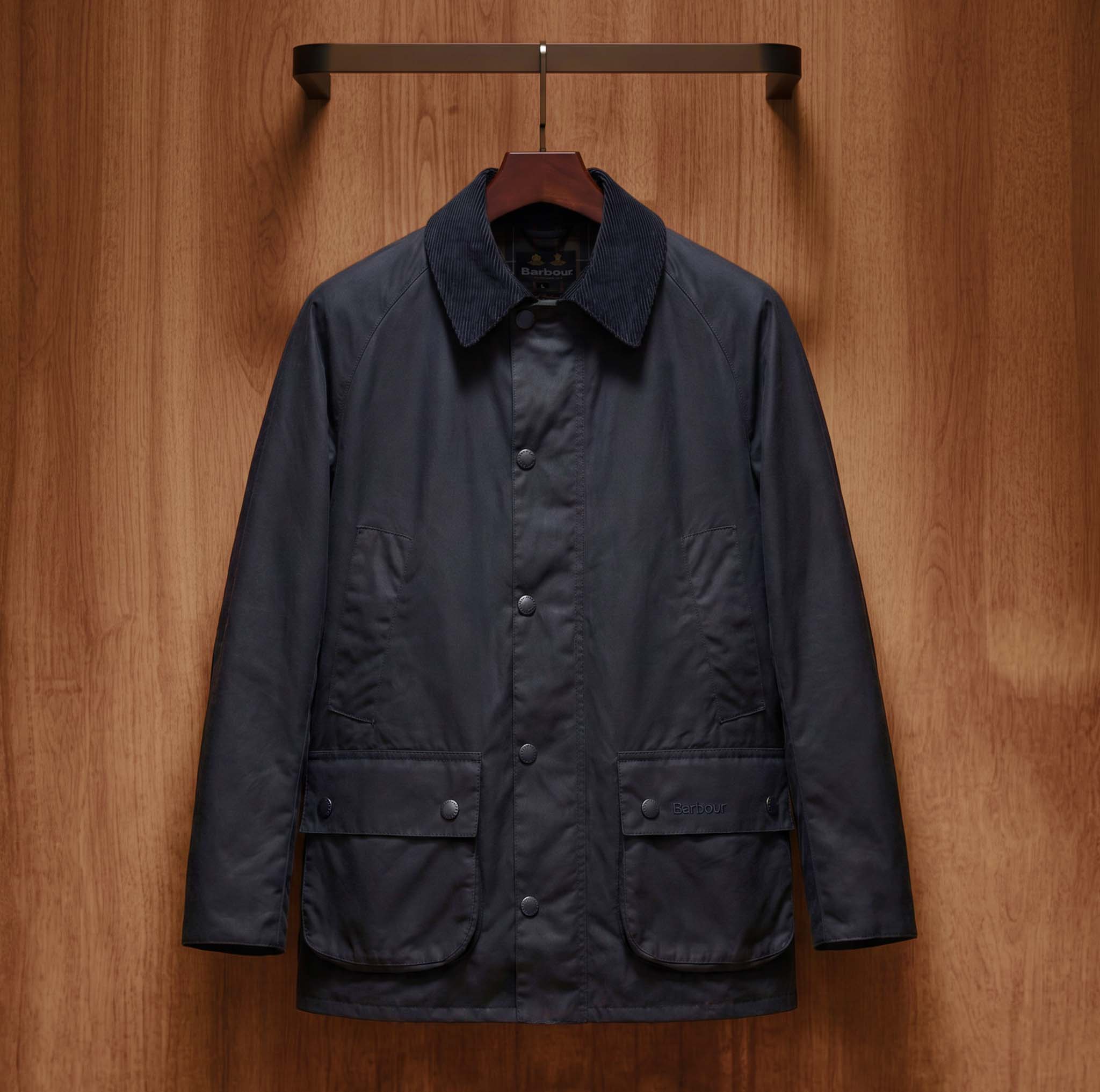 Navy blue Barbour jacket with ribbed collar and chest pockets hanging on wooden hanger against wood panelled background