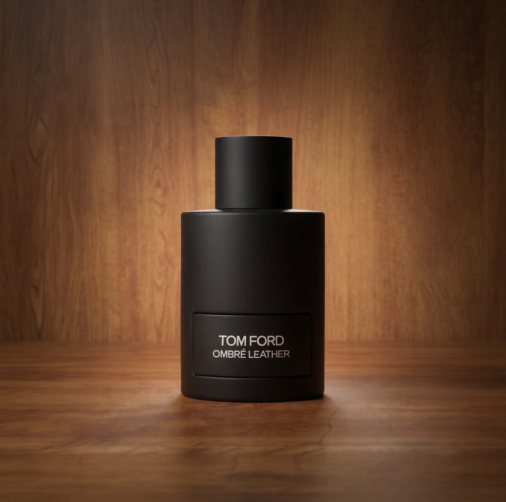 A matte black bottle of Tom Ford Ombre Leather perfume is placed on a wooden surface. The background features a softly lit wooden texture, highlighting the sleek and minimalist design of the bottle.