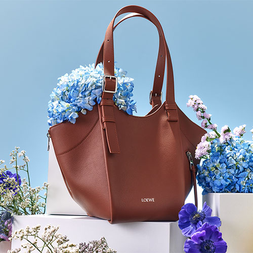 Brown leather handbag with 'LOEWE' logo, filled with blue and purple flowers, set against a light blue background.