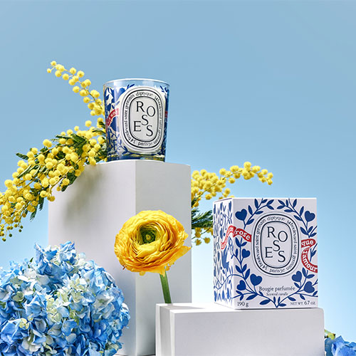 Candles labeled 'Roses' on white pedestals, surrounded by yellow flowers and blue hydrangeas against a light blue background.