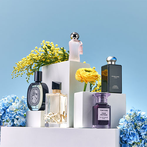Perfume bottles arranged on white platforms, surrounded by yellow and blue flowers, against a light blue background.