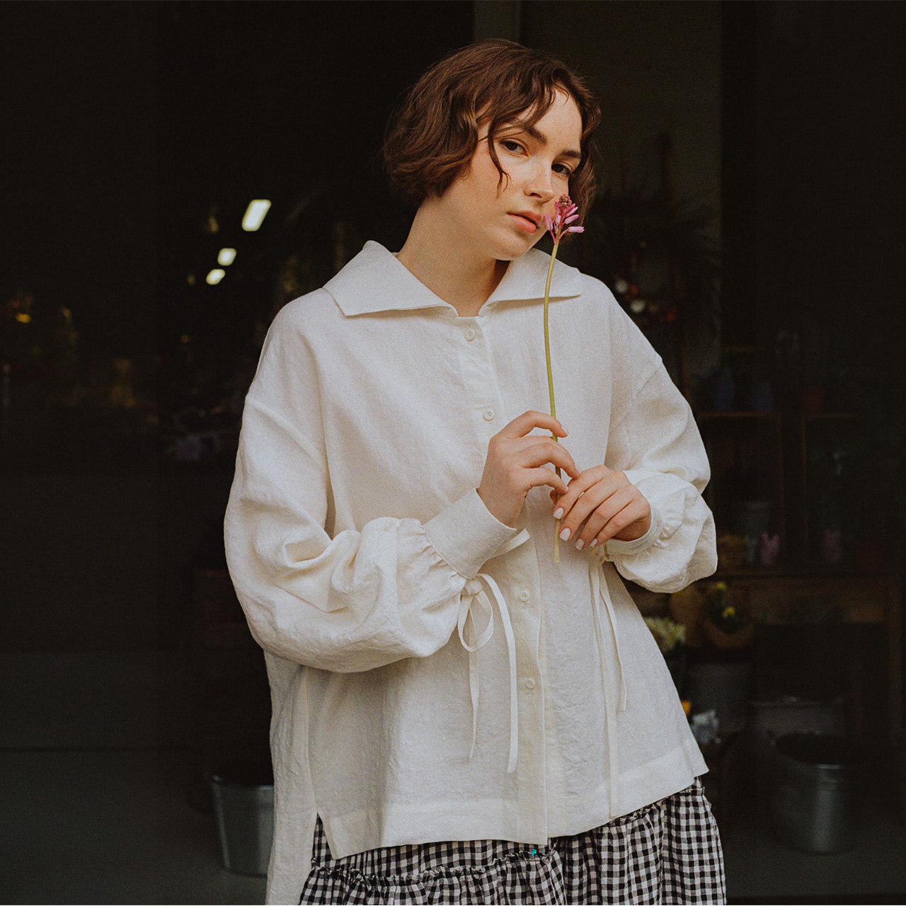 Woman wearing cream linen shirt with puffed sleeves and gingham skirt, holding a dried flower stem