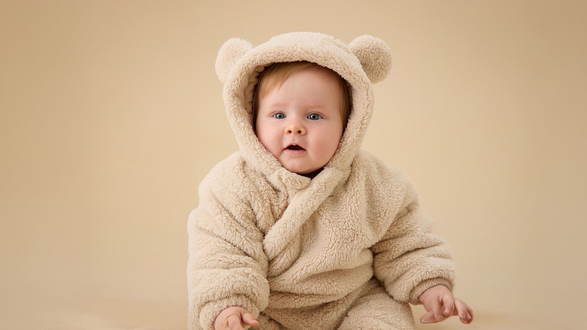 A baby in a fluffy bear costume with ears, sitting against a plain background.