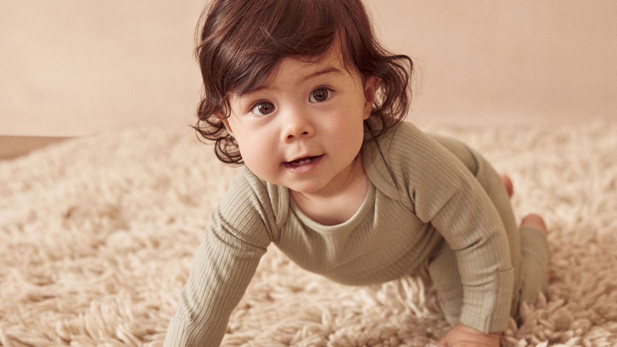 Toddler with brown curly hair wearing beige ribbed long-sleeve top, crawling on cream carpet