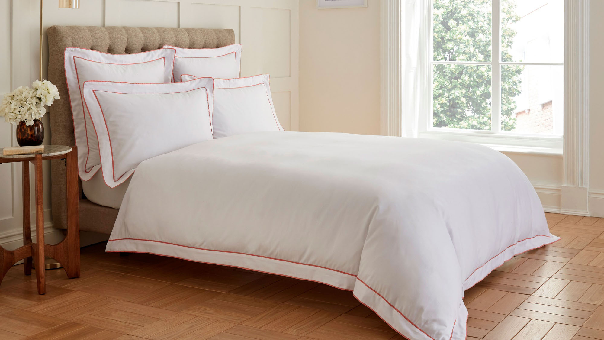 White bedding set with red trim on beige upholstered bed beside wooden side table with white flowers