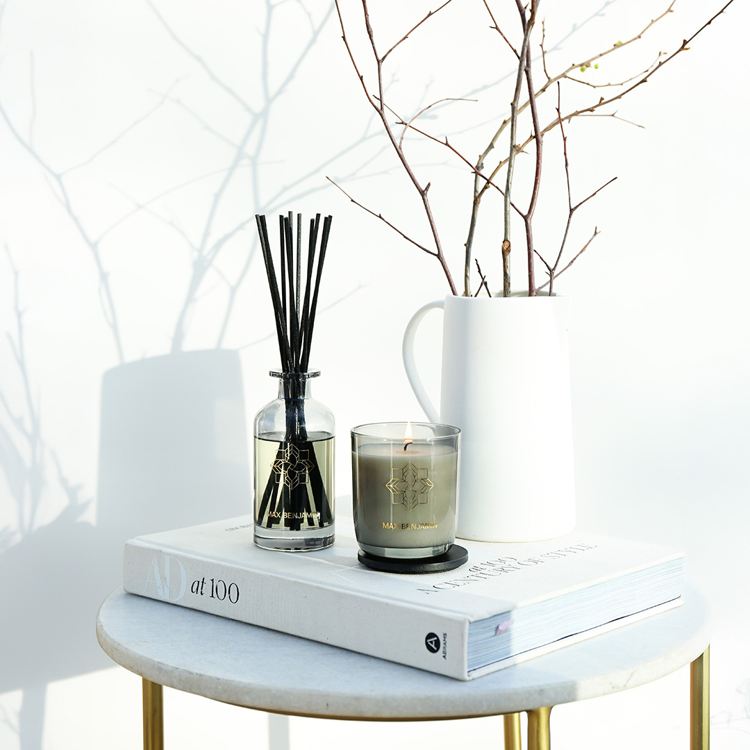 Styled tabletop with reed diffuser, scented candle, white ceramic jug with dried branches, and design book on marble table