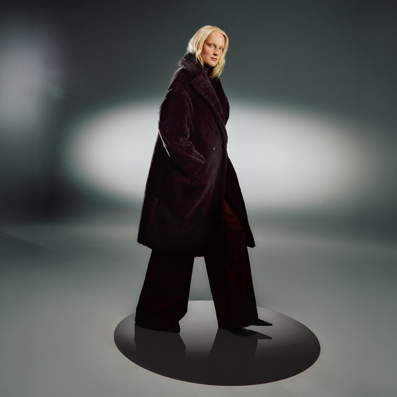 Woman with blonde hair wearing dark burgundy wool coat and black wide-leg trousers, standing against studio background