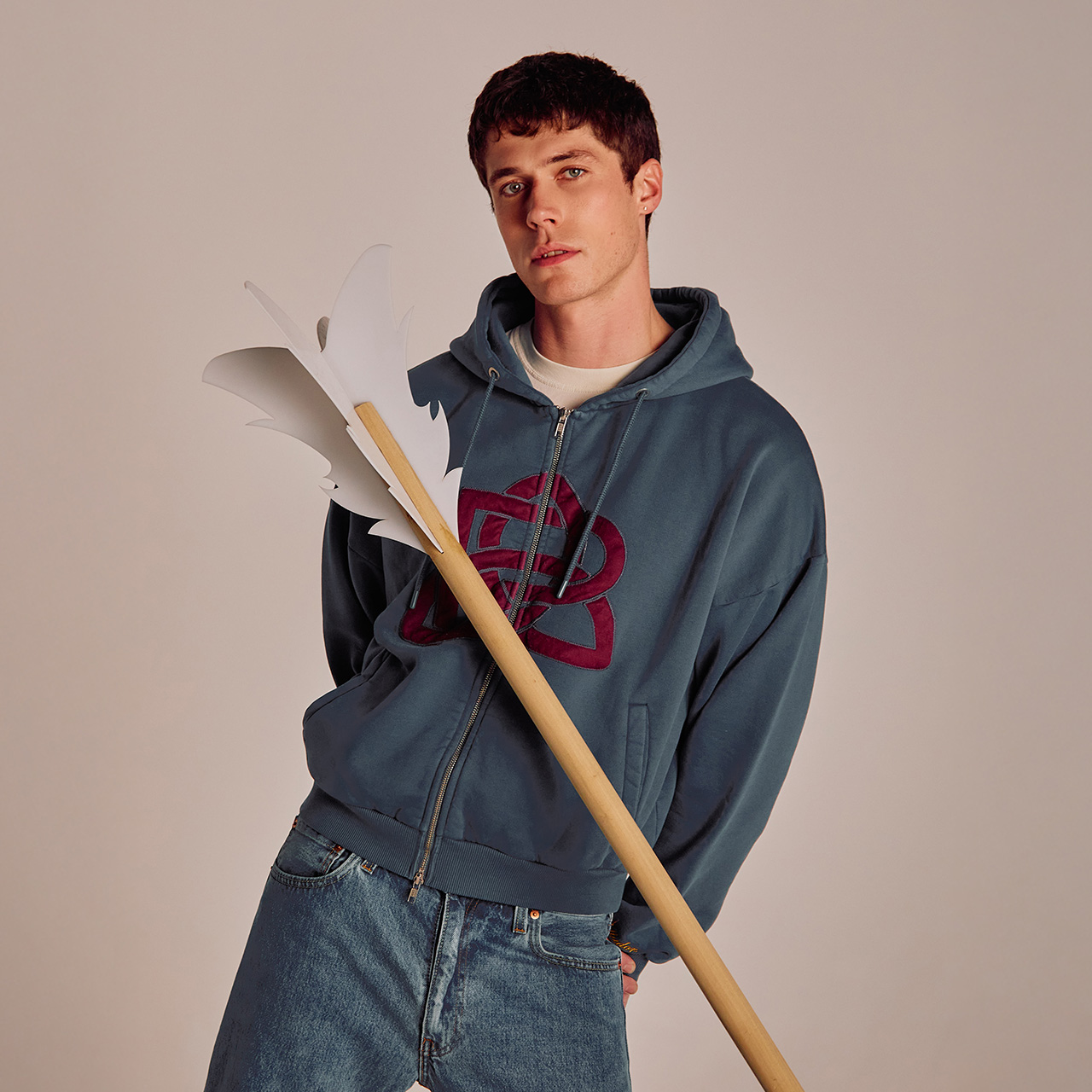 A person in a blue hoodie and jeans holds a long pole with white, stylized leaf shapes at the top.
