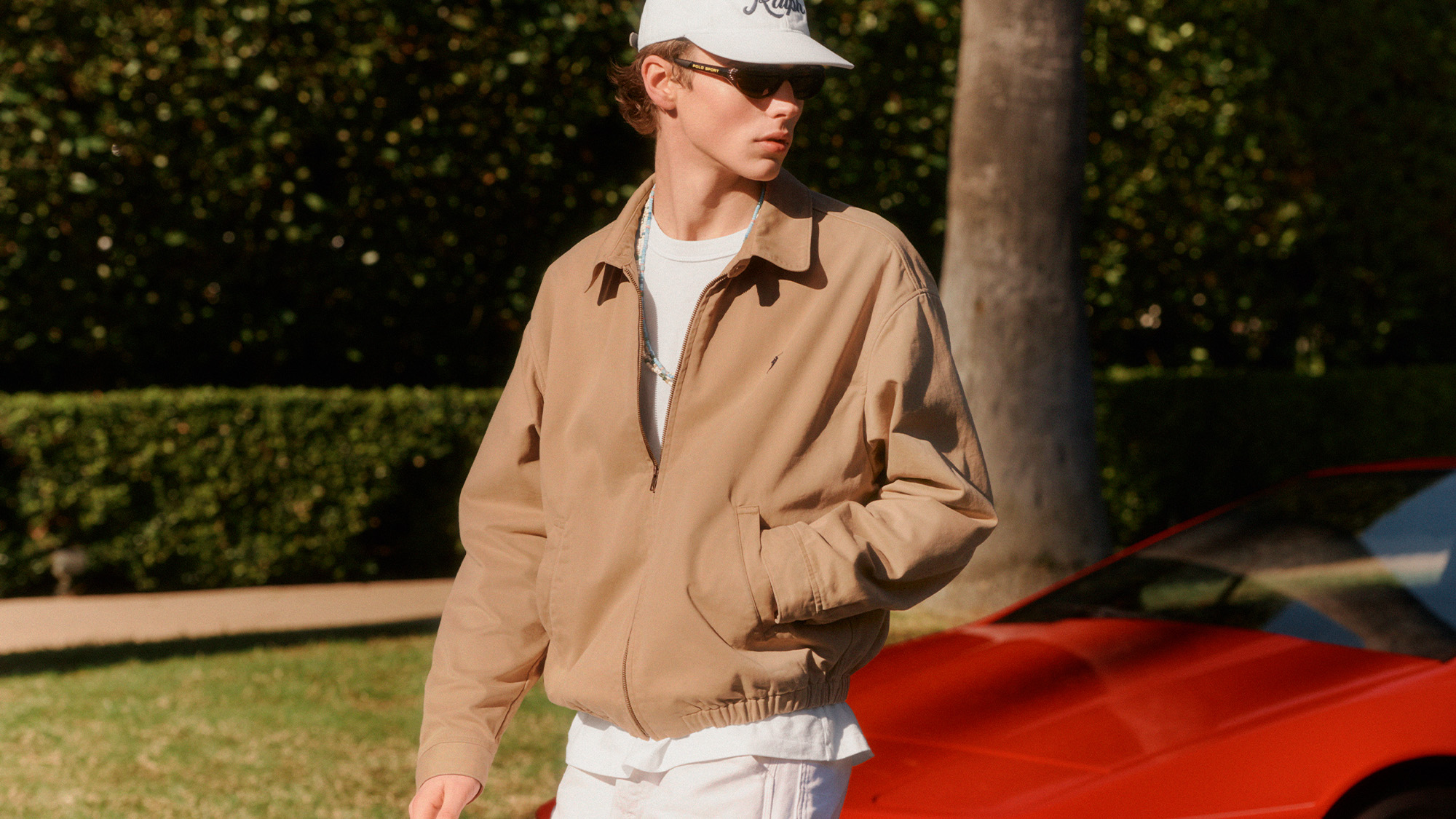 Man wearing beige jacket, white t-shirt, white cap and sunglasses against green hedge backdrop