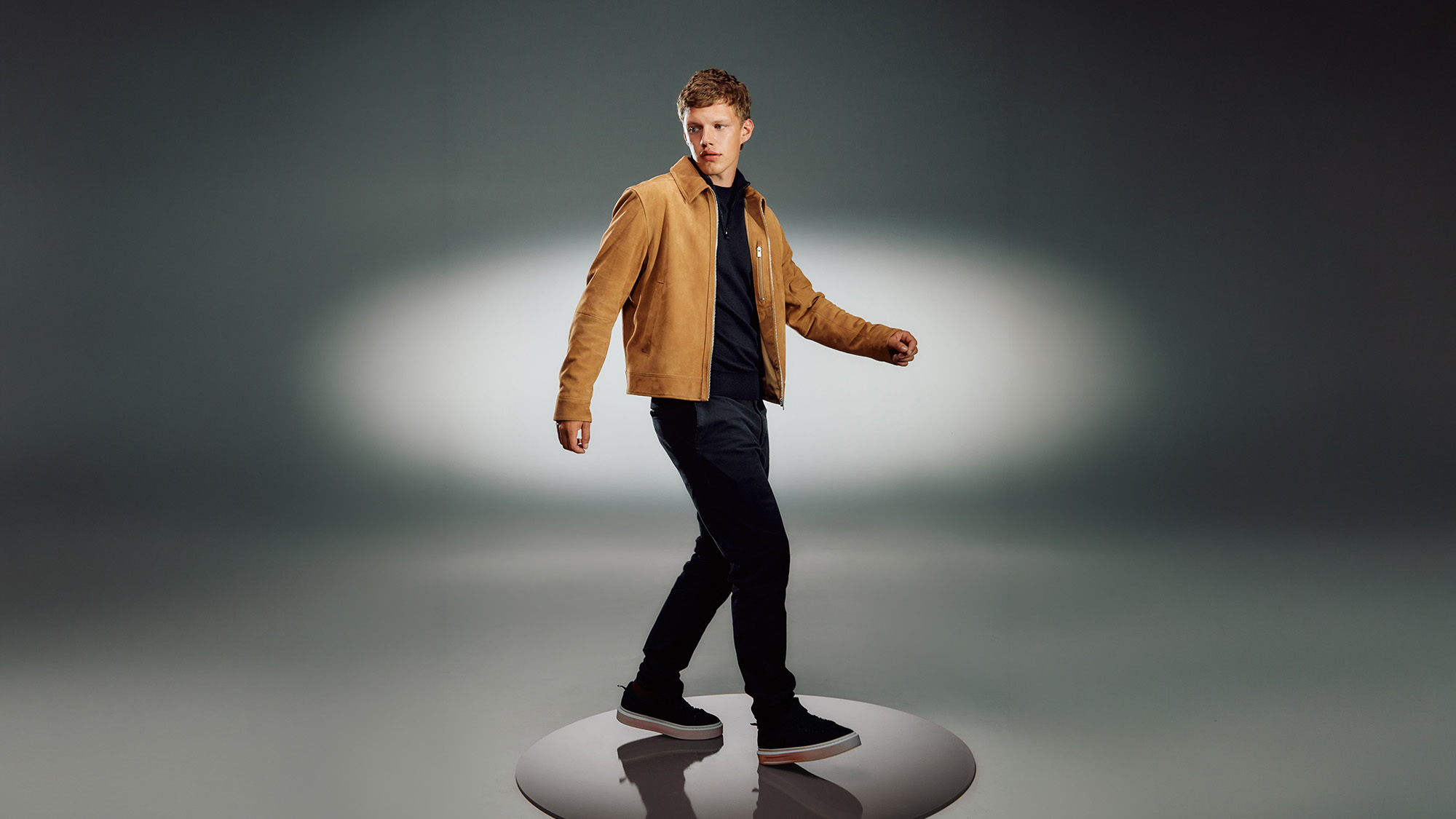 Man wearing camel harrington jacket over black jumper with navy trousers and black trainers