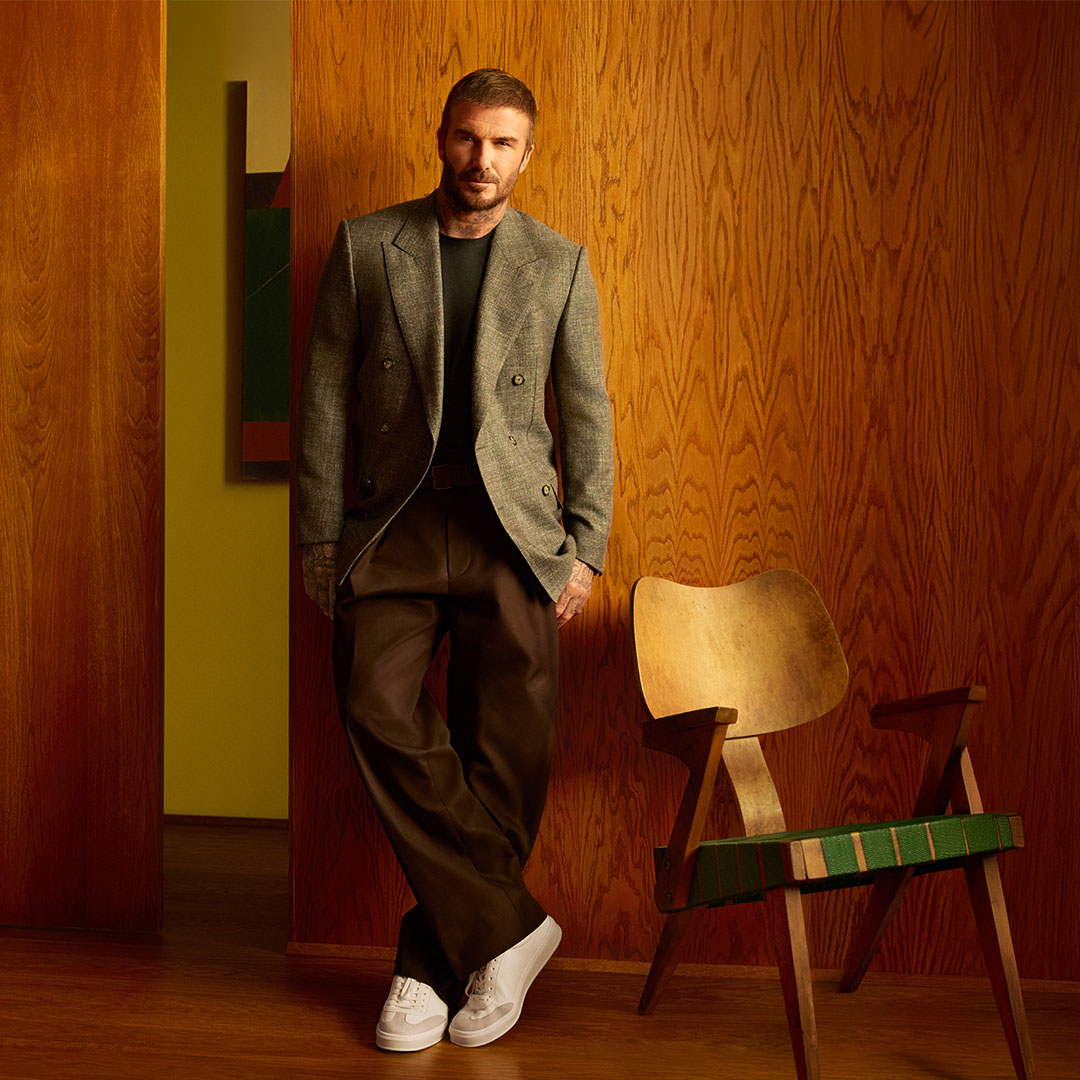 Man wearing a grey blazer and dark trousers with white trainers, standing in a wood-panelled interior