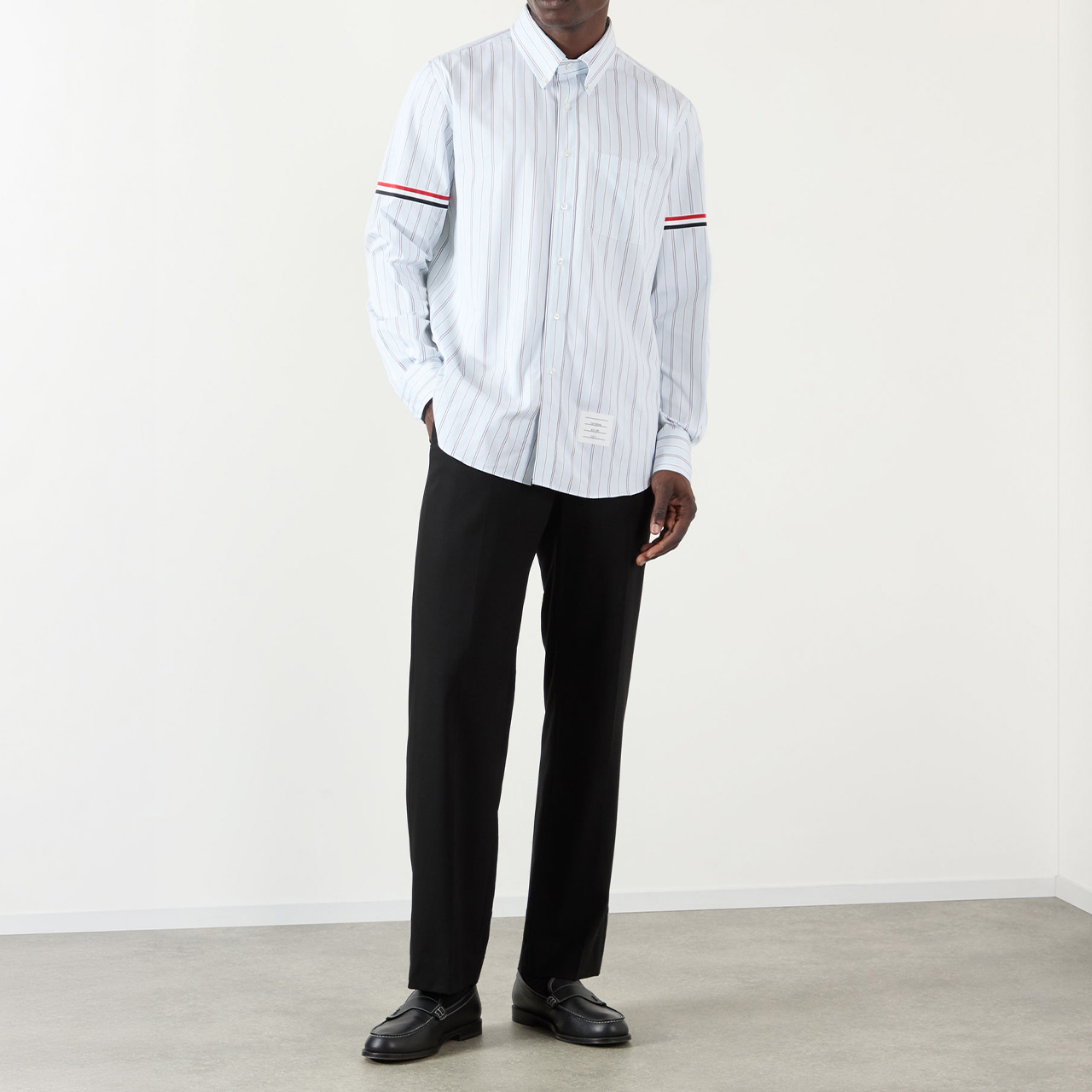 A person stands indoors wearing a light blue, striped button-up shirt with red and white bands on the sleeves. They pair it with black trousers and black shoes. The setting is minimalist with light-coloured walls and a grey floor.