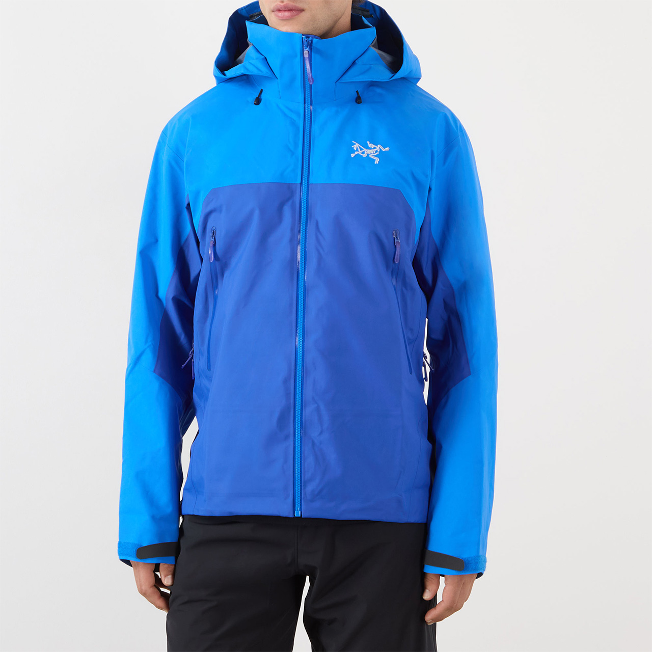 Person wearing a two-tone blue waterproof jacket with a hood and zipped pockets, featuring a logo on the chest. The jacket is paired with black trousers.