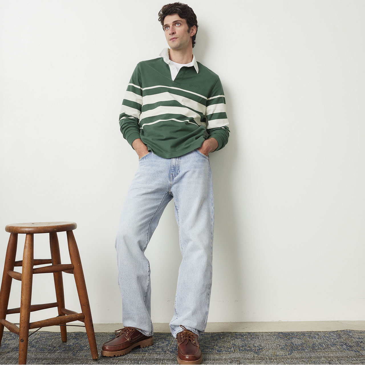 Man in a green striped sweater and jeans leans against a wall near a wooden stool.