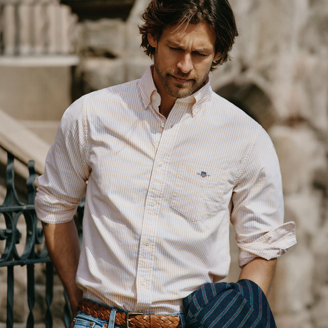 Man wearing pink and white striped shirt, brown woven belt and blue trousers against stone wall