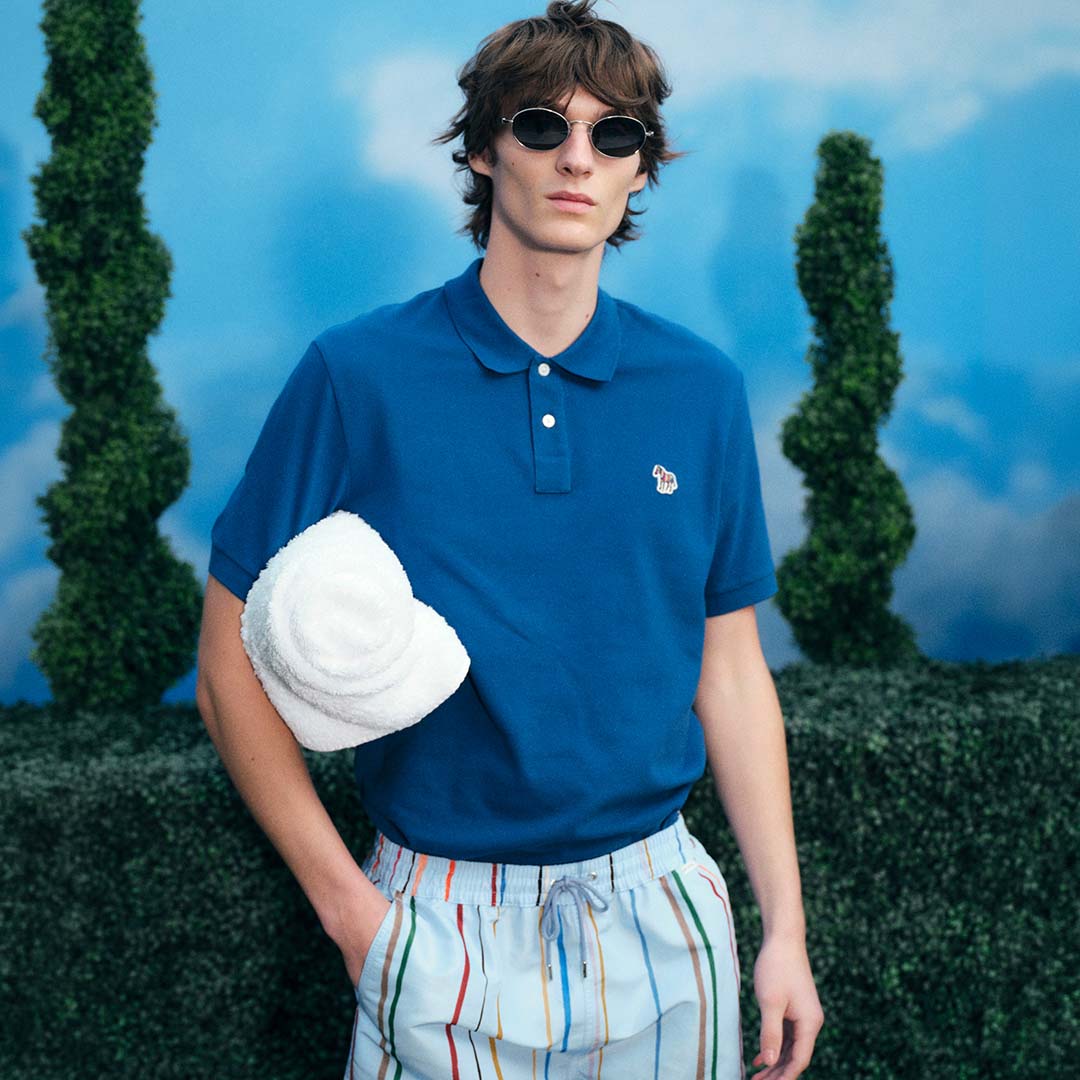Man wearing navy polo shirt, striped shorts and sunglasses holding white cap in mountain landscape setting