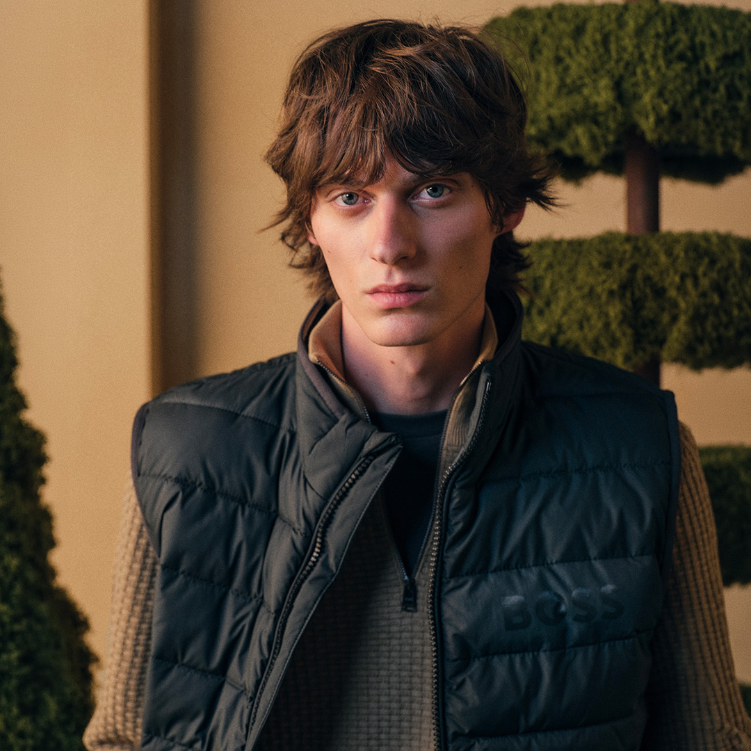 A person with medium-length hair wears a black vest and a brown sweater, standing indoors with greenery in the background.