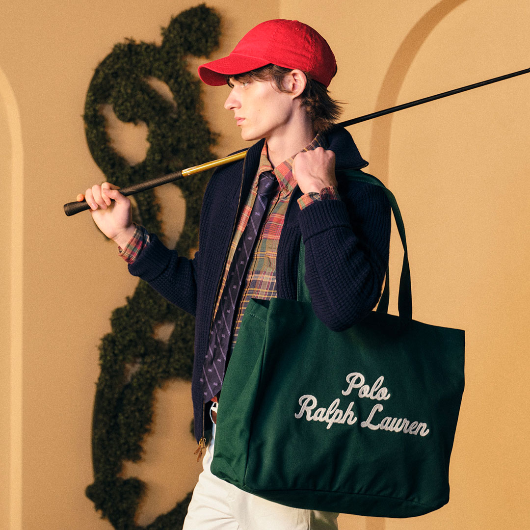 A person in a red cap holds a golf club and a green tote bag with 'Polo Ralph Lauren' text. The background features arches and greenery.