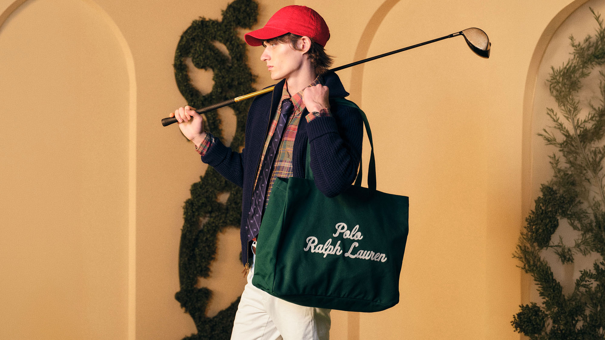 A person in a red cap holds a golf club and a green tote bag with 'Polo Ralph Lauren' text. The background features arches and greenery.