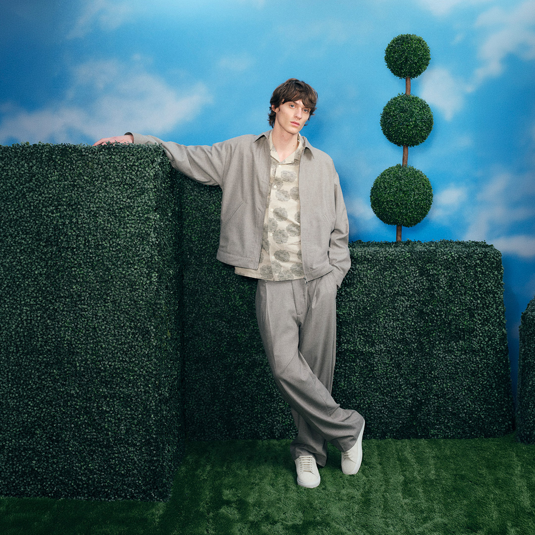 Man wearing neutral beige jacket and trousers standing against hedge maze with topiary trees, blue sky background