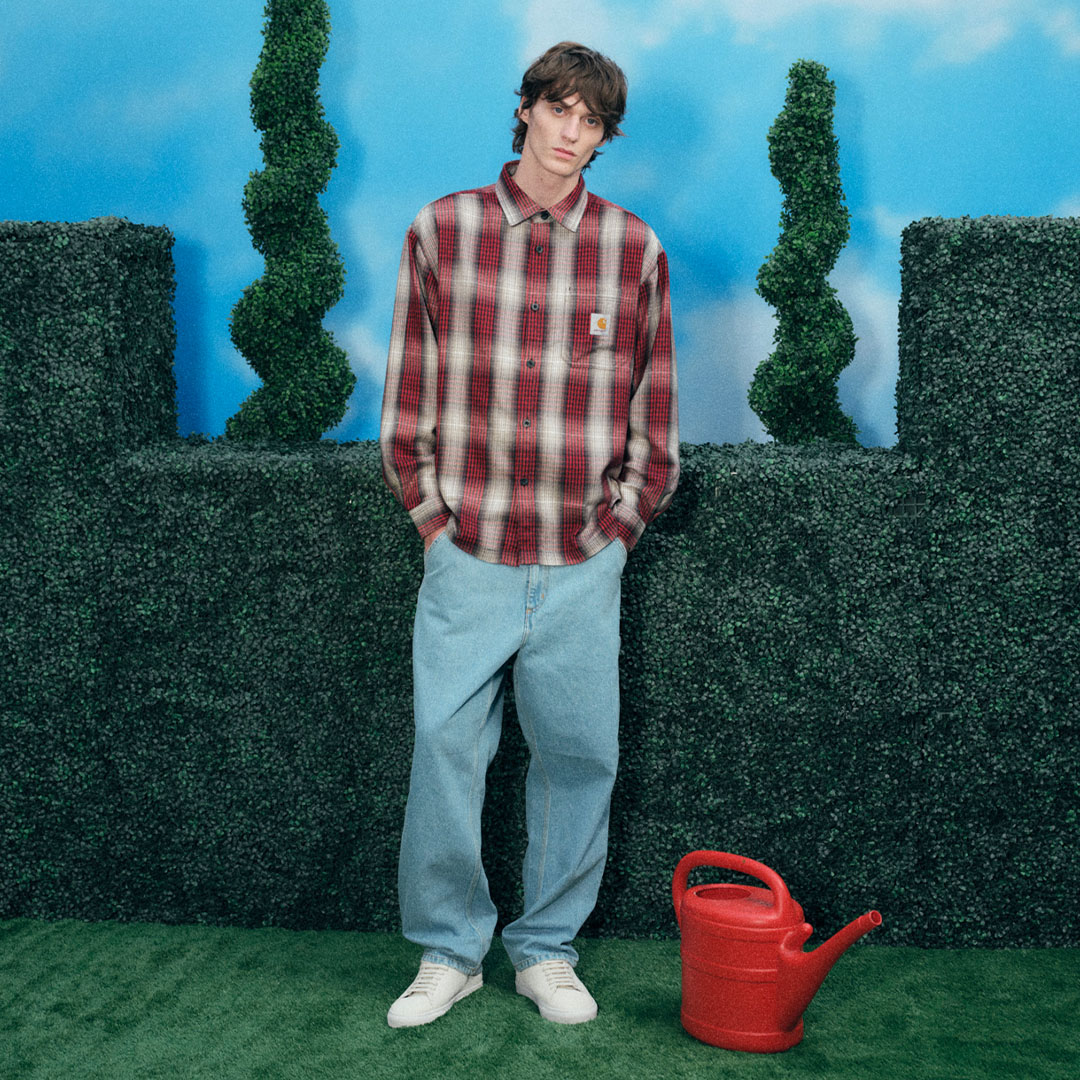 Man wearing red and white checked shirt and light blue jeans standing against green hedge backdrop