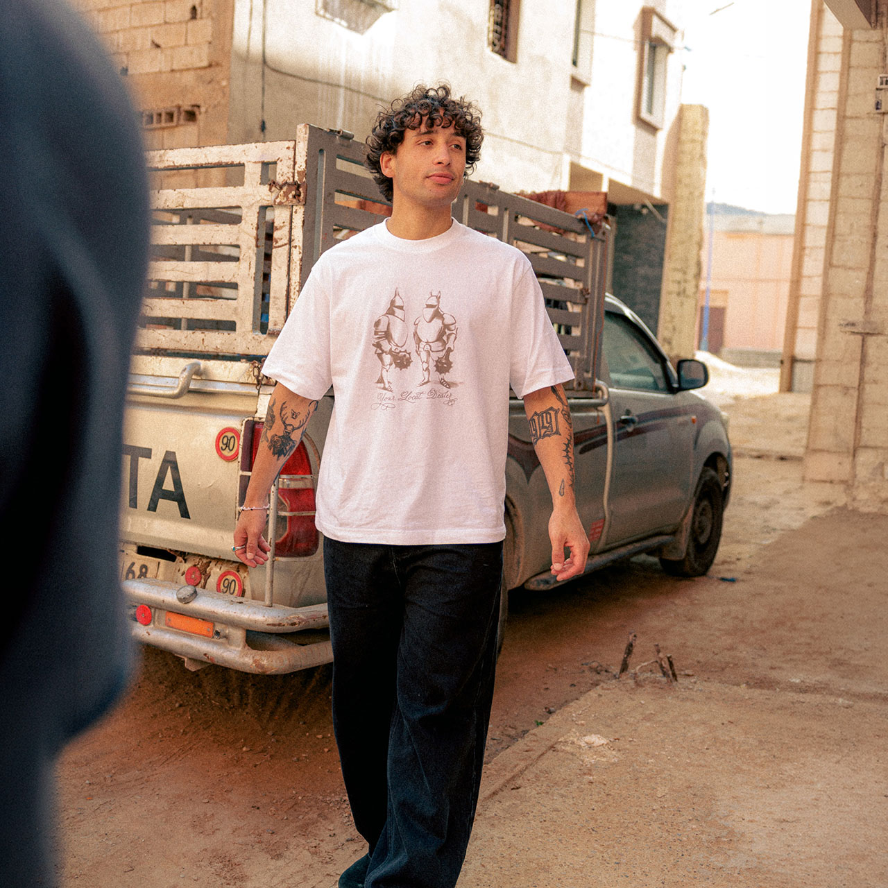 Man wearing pink graphic t-shirt and black trousers standing in industrial courtyard setting