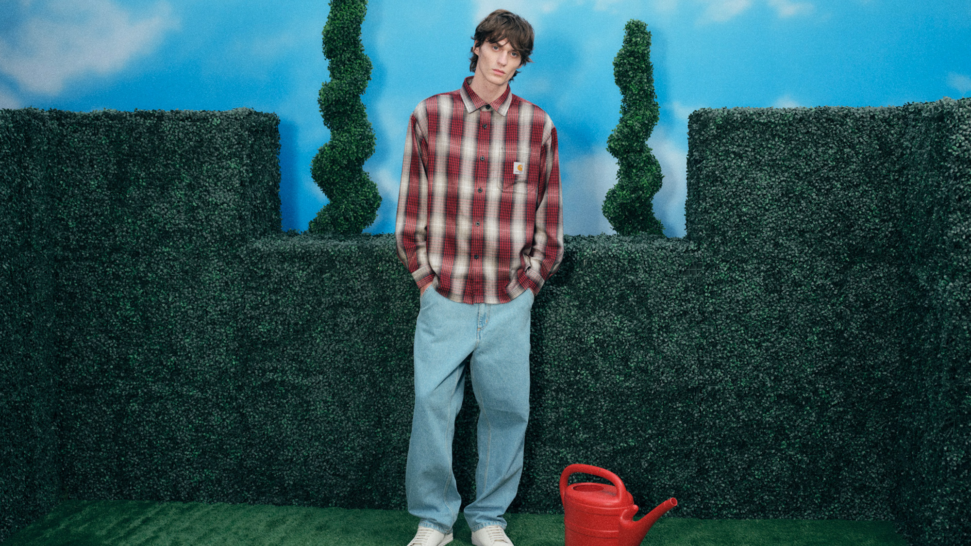 A person in a plaid shirt and jeans stands against a hedge with sculpted bushes, next to a red watering can, under a clear blue sky.