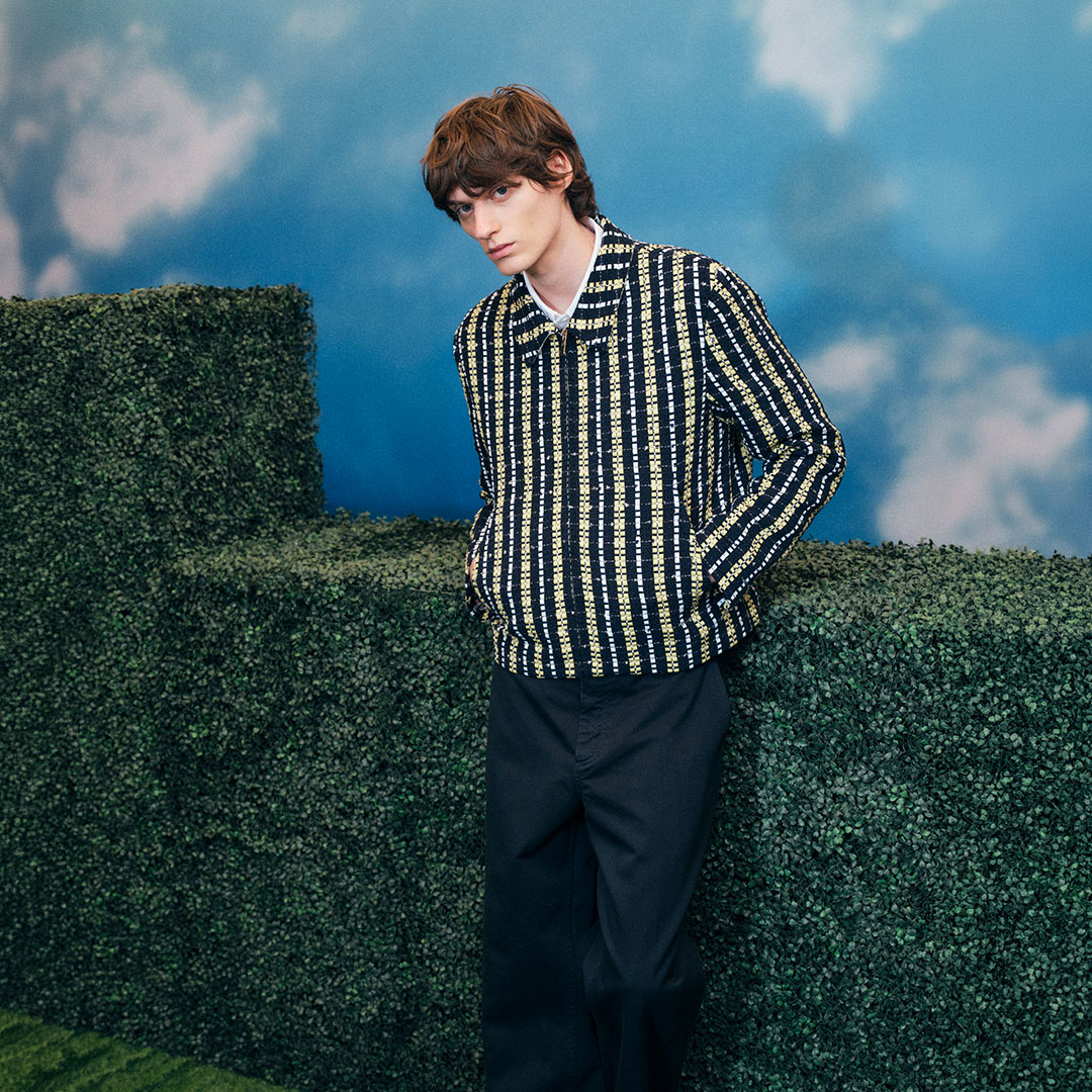 Young man wearing black and cream striped shirt and navy trousers, posed against topiary hedge
