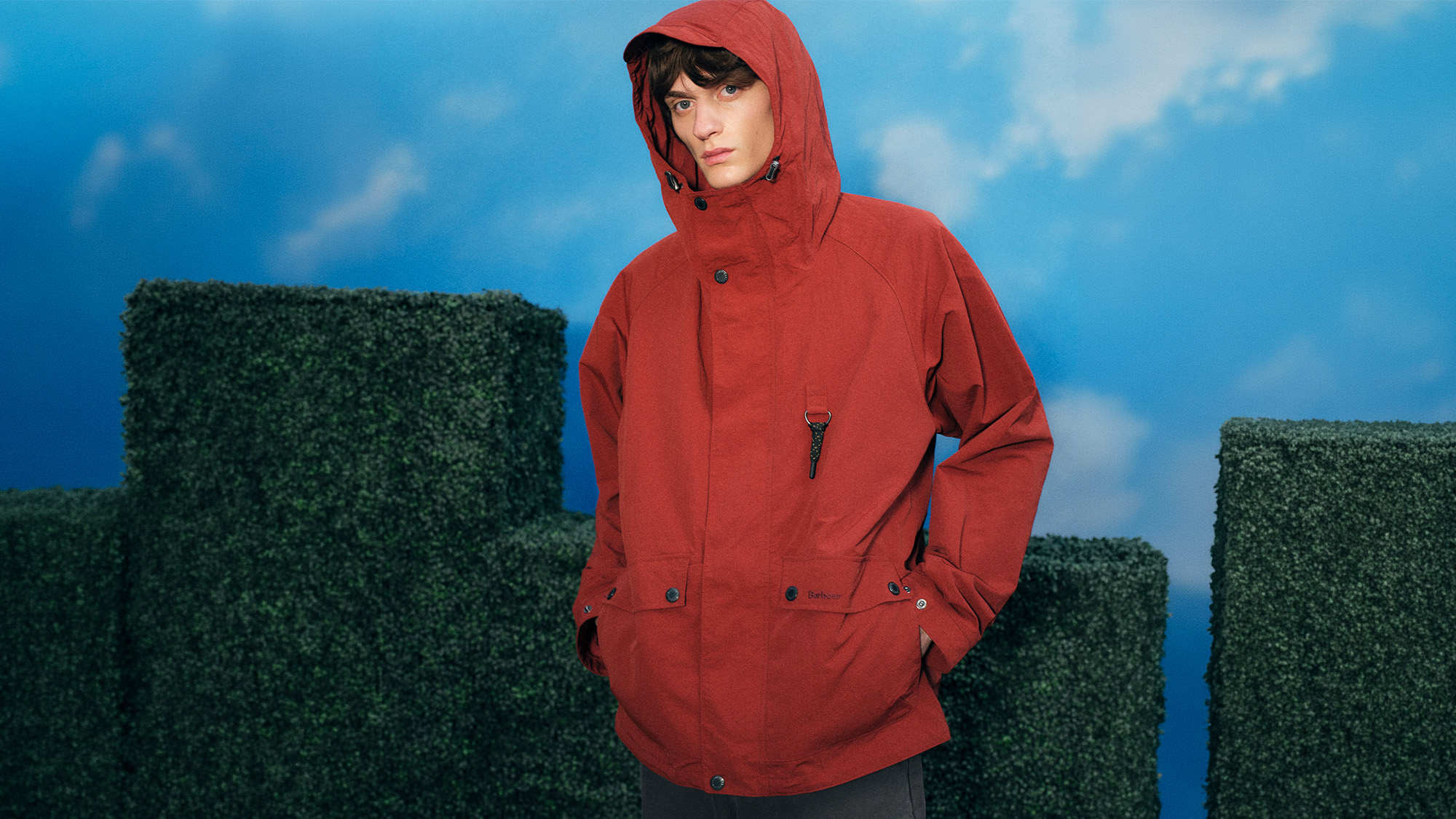 Person wearing a red hooded jacket stands against a sky backdrop with greenery.